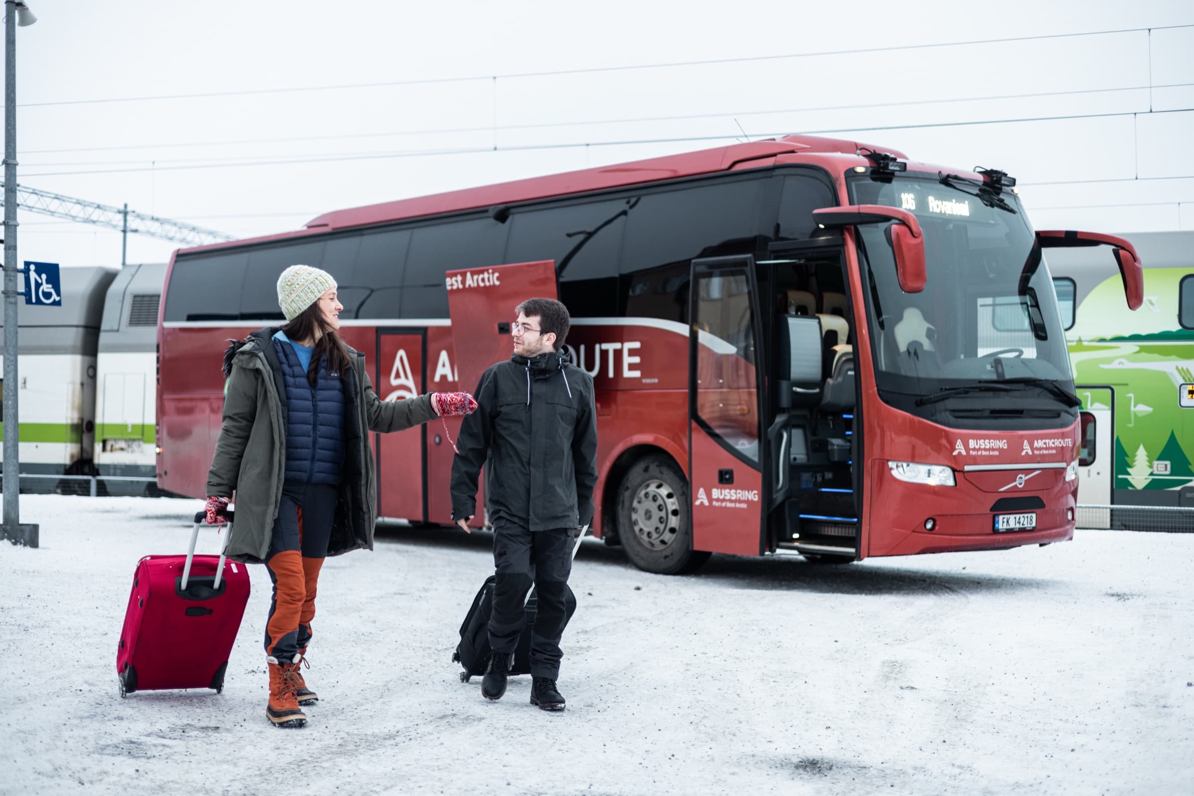 Deux voyageurs avec des bagages marchent vers un autocar rouge Arctic Route dans une gare enneigée